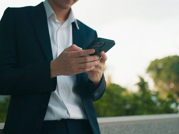 Eine Person in einem schwarzen Anzug und weißem Hemd steht an einer Mauer und schaut auf ihr Smartphone. Im Hintergrund sind unscharfe Bäume und ein blauer Himmel zu sehen.