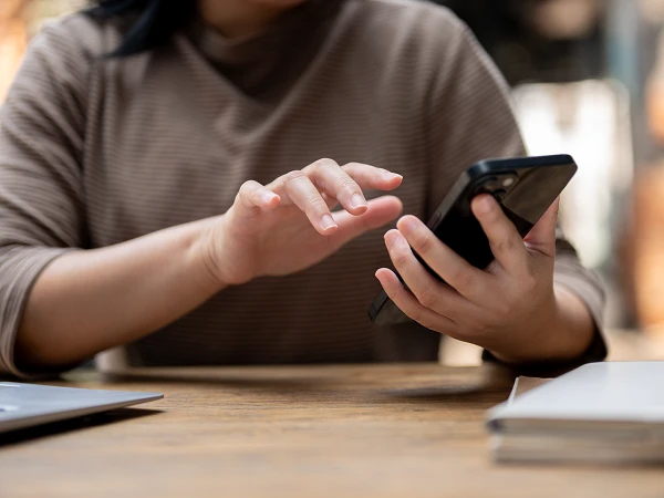 Eine Person sitzt an einem Holztisch und tippt mit einer Hand auf ihr Smartphone, während ein Laptop und ein Notizbuch in der Nähe liegen.