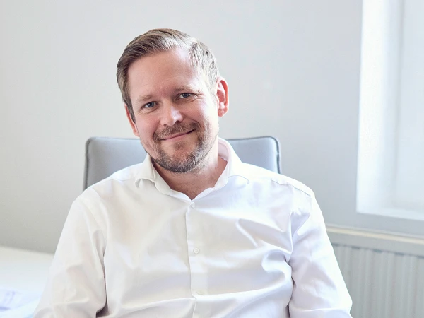 Ein Mann mit kurzem Bart und lässiger Kleidung sitzt an einem Schreibtisch in einem modernen Büro und lächelt. Auf dem Tisch liegen ein Laptop, ein Smartphone und einige Unterlagen. Im Hintergrund sind Bücher und ein Dokumentenständer zu sehen.