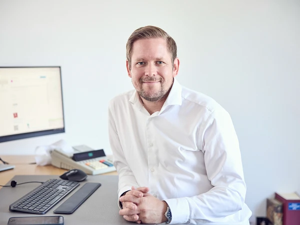 Ein Mann mit freundlichem Gesichtsausdruck sitzt an einem Schreibtisch, auf dem ein Computer und eine Tastatur stehen. Im Hintergrund ist ein Telefon und ein Regal mit Büchern sichtbar.