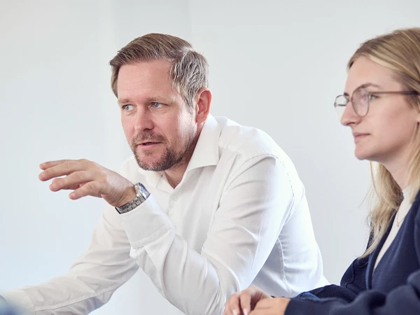 Ein Mann in einem weißen Hemd und eine Frau in einem dunklen Pullover sitzen an einem Tisch und diskutieren. Der Mann gestikuliert mit der Hand, während die Frau aufmerksam zuhört. Auf dem Tisch sind ein Laptop, ein Smartphone und verschiedene Notizmaterialien sichtbar. Im Hintergrund ist eine Weltkugel zu sehen.