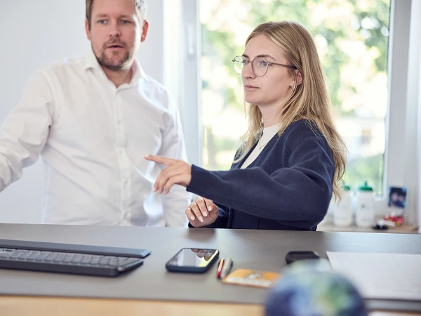 Ein Mann und eine Frau stehen an einem Schreibtisch. Die Frau zeigt mit dem Finger auf den Monitor, während der Mann aufmerksam zuschaut. Auf dem Tisch liegen ein Smartphone und andere Gegenstände. Im Hintergrund ist Licht durch ein Fenster zu sehen.