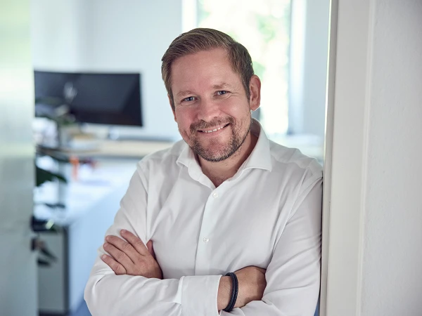 Ein lächelnder Mann mit Bart steht an einer Tür, die Arme vor der Brust verschränkt. Er trägt ein weißes Hemd und wirkt freundlich. Im Hintergrund sind unscharf Büroeinrichtungen und ein Monitor zu erkennen.