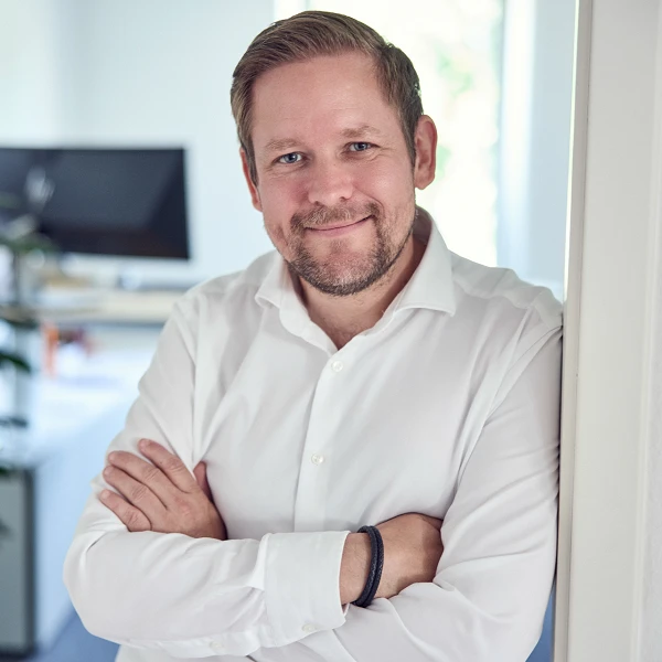 Ein Mann mit weißem Hemd steht lächelnd an einer Tür und hat die Arme vor der Brust verschränkt. Im Hintergrund ist ein Büro mit einem Monitor und Pflanzen sichtbar.