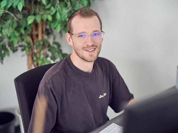 Ein Mann mit Brille und einem schwarzen T-Shirt sitzt an einem Schreibtisch und lächelt, während er auf einen Computerbildschirm schaut. Im Hintergrund sind grüne Pflanzen und ein Tisch mit einem kleinen Objekt zu sehen.