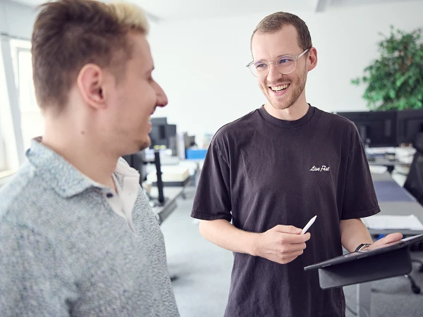 Zwei Männer in einem modernen Büro sprechen miteinander; einer von ihnen hält ein Tablet und einen Stift und lächelt, während der andere interessiert zuhört. Hintergrund mit Schreibtischen und Pflanzen sichtbar.