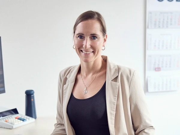 Eine Frau in einem modernen Büro steht lächelnd an einem Schreibtisch, der mit einem Computer und Bürobedarf ausgestattet ist. Sie trägt ein schwarzes Outfit und eine beige Blazerjacke. Im Hintergrund sind Kalender und weitere Bürodetails sichtbar.