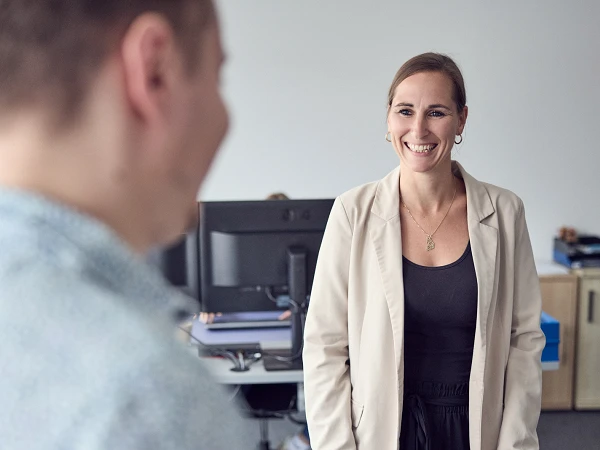 Eine Frau mit langen, braunen Haaren und einem blauen Blazer lächelt, während sie an einem Tisch in einem modernen Büro steht, vor einem Mann, der ihr zugewandt ist. Im Hintergrund sind Büroeinrichtungen und ein Computer sichtbar.