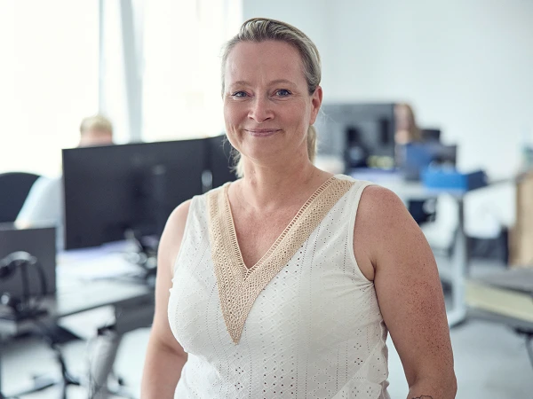 Eine Frau mit blonden Haaren steht in einem modernen Büro, lächelt und trägt ein weißes, ärmelloses Oberteil. Im Hintergrund sind Schreibtische mit Computern zu sehen.