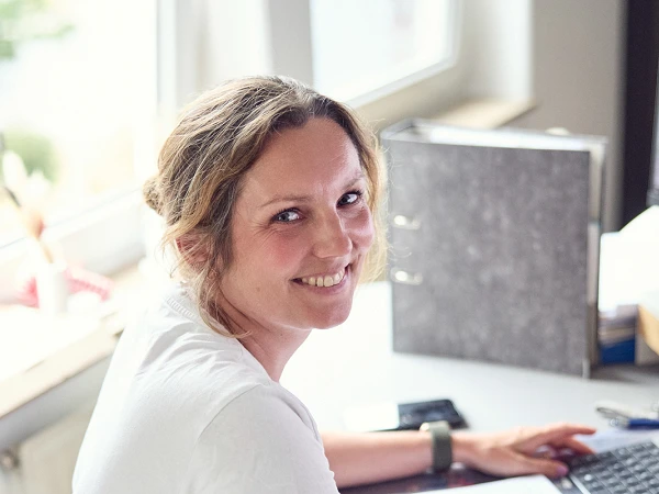 Eine Frau mit lässiger Kleidung sitzt an einem Schreibtisch und lächelt, während sie an ihrem Computer arbeitet. Im Hintergrund ist eine zweite Person zu sehen, die ihr zugewandt ist. Auf dem Schreibtisch liegen Unterlagen und es gibt eine helle, freundliche Atmosphäre durch das einfallende Licht.