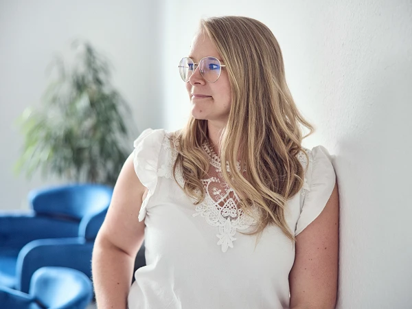 Eine Frau mit blonden, lockigen Haaren und einer weißen Bluse mit Spitze steht lächelnd an einer Wand und schaut nachdenklich zur Seite. Im Hintergrund sind blau gepolsterte Stühle und eine Pflanze zu sehen.