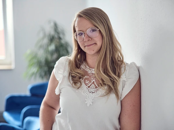Eine Person mit langen, blonden Haaren und runden Brillengestellen lehnt an einer Wand und lächelt in die Kamera. Im Hintergrund sind eine Pflanze und blaue Sitzmöbel zu sehen.