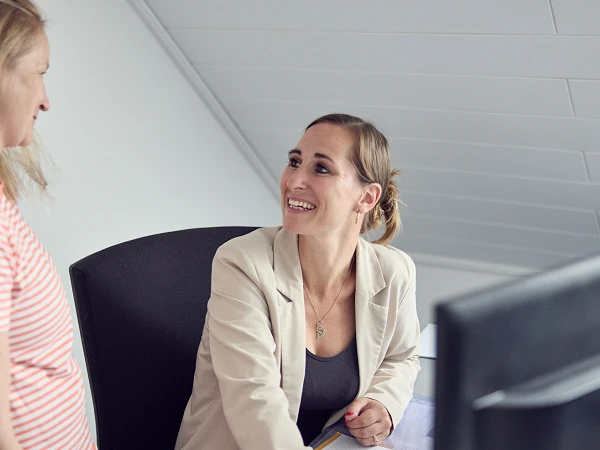 Eine Frau im blauen Blazer sitzt an einem Schreibtisch und spricht lächelnd mit einer Frau in gestreiftem Shirt, die neben ihr steht. Am Schreibtisch liegen Papierunterlagen und ein Stift. Im Hintergrund ist ein Computer sichtbar.