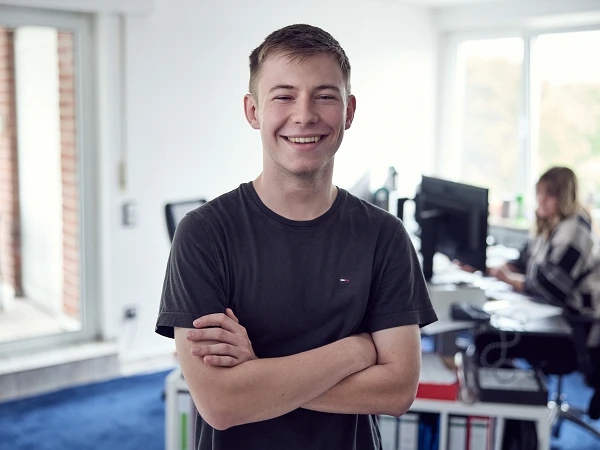 Ein junger Mann mit einem breiten Lächeln und überkreuzten Armen steht in einem modernen Büro, während im Hintergrund eine andere Person am Schreibtisch arbeitet.