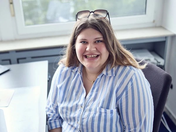 Eine lächelnde Frau mit langen, braunen Haaren sitzt an einem Schreibtisch in einem Büro, umgeben von Computerbildschirmen und Büromaterialien. Sie trägt ein gestreiftes Oberteil und hat eine Sonnenbrille im Haar. Im Hintergrund sind Fenster mit Blick ins Freie zu sehen.