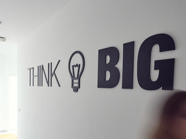 Ein langen Flur mit einem blauen Teppich, an der Wand steht in großen Buchstaben „THINK BIG“. Eine Person bewegt sich schnell durch den Flur und ist unscharf abgebildet.