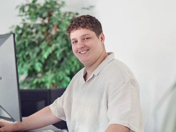 Ein junger Mann mit lockigem Haar sitzt an einem Schreibtisch vor einem Computer und lächelt in die Kamera. Im Hintergrund ist eine grüne Pflanze zu sehen. Auf dem Tisch liegen einige Unterlagen und ein Ordner.