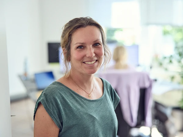 Eine lächelnde Frau steht in einem hellen Büro, während im Hintergrund eine andere Person an einem Computer arbeitet. Die Frau trägt ein dunkelgrünes T-Shirt und hat lockeres Haar.