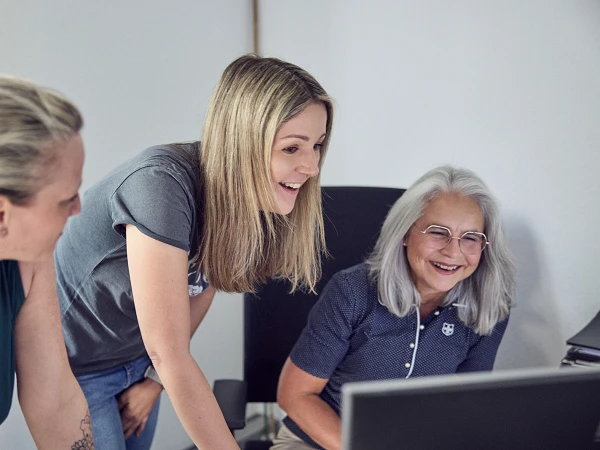 Drei Frauen stehen um einen Schreibtisch und schauen auf einen Computerbildschirm, während sie begeistert miteinander sprechen. Die Frau in der Mitte hat längere, blonde Haare und lächelt, während die anderen beiden Frauen, eine mit kurzem, grauen Haar und die andere mit Schulterlangen Haaren, ebenfalls interessiert sehen.