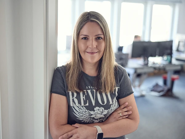 Eine lächelnde Frau mit langen, blonden Haaren steht entspannt an einer Wand in einem Büro, während sie einen grauen T-Shirt mit dem Schriftzug "REVOLT" trägt. Im Hintergrund sind Schreibtische und Computer sichtbar.
