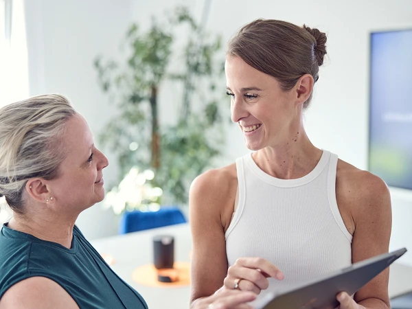 Zwei Frauen stehen in einem modernen Büro und schauen auf ein Tablet. Eine der Frauen trägt ein ärmelloses, weißes Oberteil und lächelt, während die andere in einem grünen Oberteil ebenfalls lächelt und auf den Bildschirm zeigt. Im Hintergrund sind Pflanzen und ein Bildschirm zu sehen.