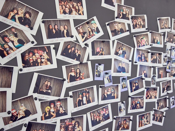 Eine Wand mit vielen polaroidartigen Fotos, die verschiedene Gruppen von Menschen bei Feiern oder Zusammenkünften zeigen, sind mit Klammern befestigt.