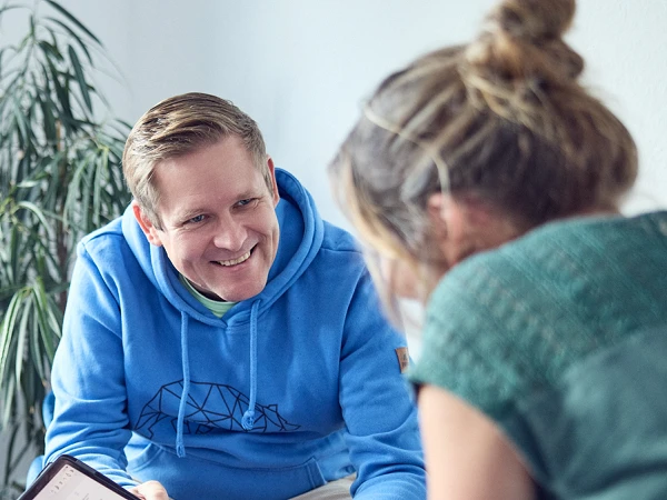 Drei Personen sitzen in einem hellen Raum auf blauen Sesseln um einen runden Tisch. Ein Mann in einem blauen Hoodie zeigt einer Frau etwas auf einem Tablet, während die andere Frau ihm interessiert zuschaut. Auf dem Tisch stehen eine rote und eine weiße Tasse. Im Hintergrund ist eine Pflanze zu sehen.