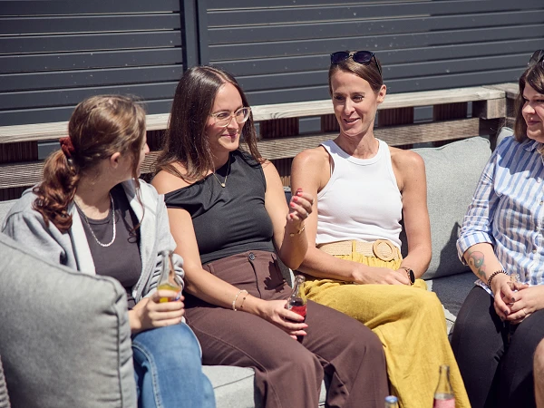 Fünf Frauen sitzen auf einer modernen Couch im Freien, trinken Erfrischungsgetränke und unterhalten sich angeregt. Eine Frau trägt ein schwarzes Oberteil, eine andere ein weißes Top und eine hellbraune Hose, während eine dritte in einem gelben Rock gekleidet ist. Die Szene strahlt eine entspannte, gesellige Atmosphäre aus.