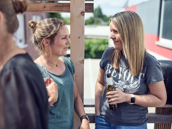 Zwei Frauen unterhalten sich auf einer sonnigen Terrasse, während sie Getränke in der Hand halten. Im Hintergrund sind unscharfe Silhouetten von weiteren Personen zu sehen und die Landschaft ist grün.