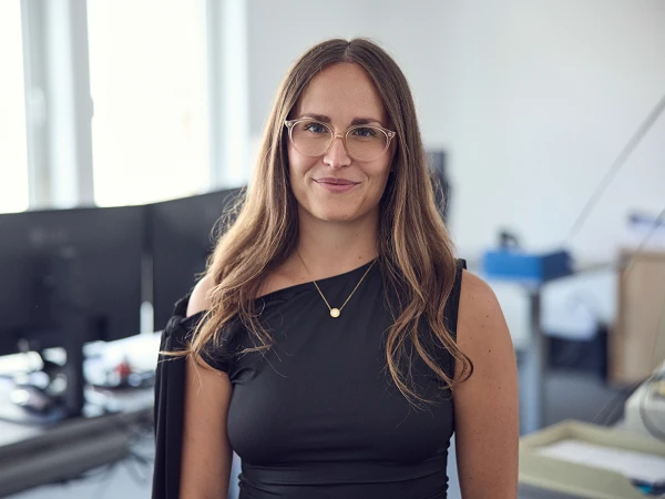 Eine Frau mit langen, braunen Haaren und einer schwarzen, schulterfreuen Bluse lächelt in die Kamera. Sie trägt eine Kette mit Anhänger und steht in einem modernen Büro, wo mehrere Computer im Hintergrund zu sehen sind.