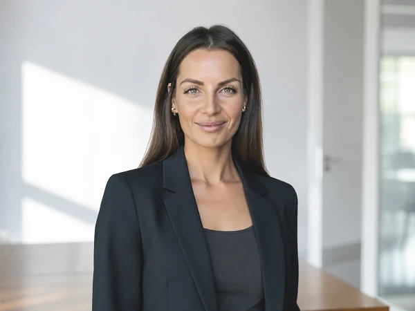 Eine Frau mit langen, braunen Haaren trägt einen schwarzen Blazer und steht in einem modernen, hellen Raum mit großen Fenstern. Sie lächelt leicht und hat einen selbstbewussten Ausdruck.