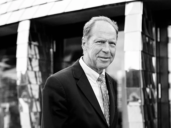 Eine Personen steht in einem eleganten Anzug und lächelt, während sie vor einem modernen, glänzenden Gebäude mit schrägem Dach posiert.